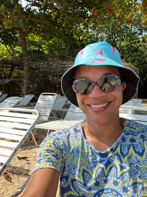 African American woman in sunglasses, blue and yellow paisley rash shirt and light blue bucket hat with flamingos on it sitting on white lounge chair under tropical trees